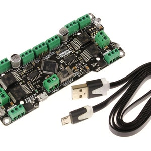Puede incluir: Una placa de circuito negro con conectores verdes, etiquetada como "XMOTION MEC", y un cable USB. La placa tiene varios componentes electrónicos y el texto "Start: D11". El cable USB es negro con conectores blancos.