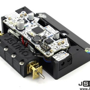 Può includere: Un telaio robotico in plastica nera dotato di una scheda a circuito bianco etichettata JSMOTION, due motori color ottone con alberi metallici e un supporto motore nero con il testo JSUMO impresso in rilievo.