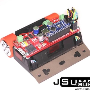 Può includere: Un telaio per robot sumo con una scheda a circuito stampato rossa, un microcontrollore blu e due ruote arancioni. Il dispositivo include una lama metallica nera sul davanti, morsettiere verdi, condensatori argentati e cavi multicolore.