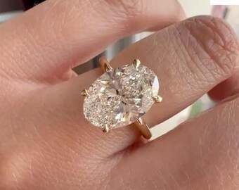 May include: A gold ring with a large oval-shaped diamond. The diamond is set in a prong setting and has a brilliant cut. The ring is on a finger.