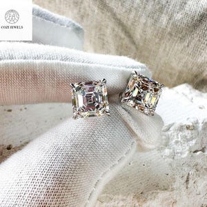 May include: A pair of square-cut diamond stud earrings. The earrings are set in a silver metal and are displayed on a white cloth. The earrings are from Cozi Jewels.