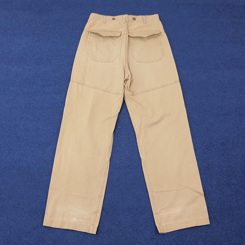 May include: Khaki-colored pants with two back pockets and a button closure. The pants have a wide leg design and are made of a durable fabric. The stitching is visible, and the pants are displayed against a blue background.