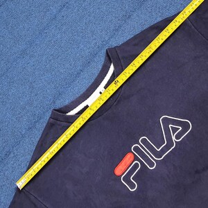 Puede incluir: Sudadera azul marino de cuello redondo con el logo de FILA en blanco y rojo. La sudadera tiene un cuello blanco y se mide con una cinta m&eacute;trica amarilla. La cinta m&eacute;trica muestra una longitud de aproximadamente 53 cm.