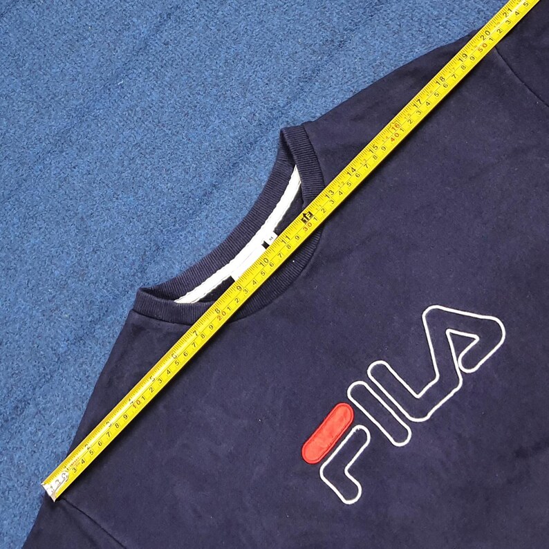 Puede incluir: Sudadera azul marino de cuello redondo con el logo de FILA en blanco y rojo. La sudadera tiene un cuello blanco y se mide con una cinta m&eacute;trica amarilla. La cinta m&eacute;trica muestra una longitud de aproximadamente 53 cm.