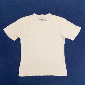 Puede incluir: Camiseta blanca de manga corta con una firma negra "&copy; K. Haring" impresa en la espalda.