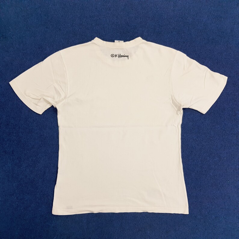 Puede incluir: Camiseta blanca de manga corta con una firma negra "&copy; K. Haring" impresa en la espalda.