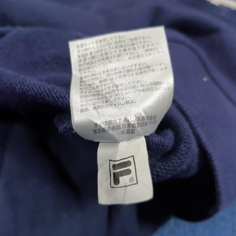 Puede incluir: Una etiqueta de prenda azul marino con texto japon&eacute;s y un logotipo blanco y negro. La etiqueta est&aacute; adherida a una tela azul marino. El logotipo es un cuadrado blanco con una "F" y una "L" negras en su interior.