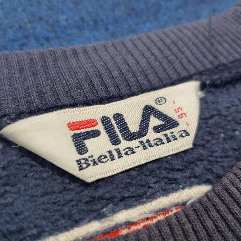Puede incluir: Primer plano de una sudadera azul con una etiqueta blanca. La etiqueta presenta el logotipo de FILA en rojo y azul, con el texto "Biella-Italia" debajo. La sudadera tiene un cuello acanalado.