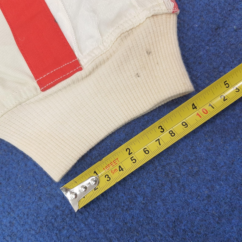 Puede incluir: Primer plano de una tela color crema con una franja roja y un pu&ntilde;o acanalado. Una cinta m&eacute;trica amarilla se coloca diagonalmente sobre la imagen, mostrando medidas en cent&iacute;metros. La tela parece ser parte de una prenda.