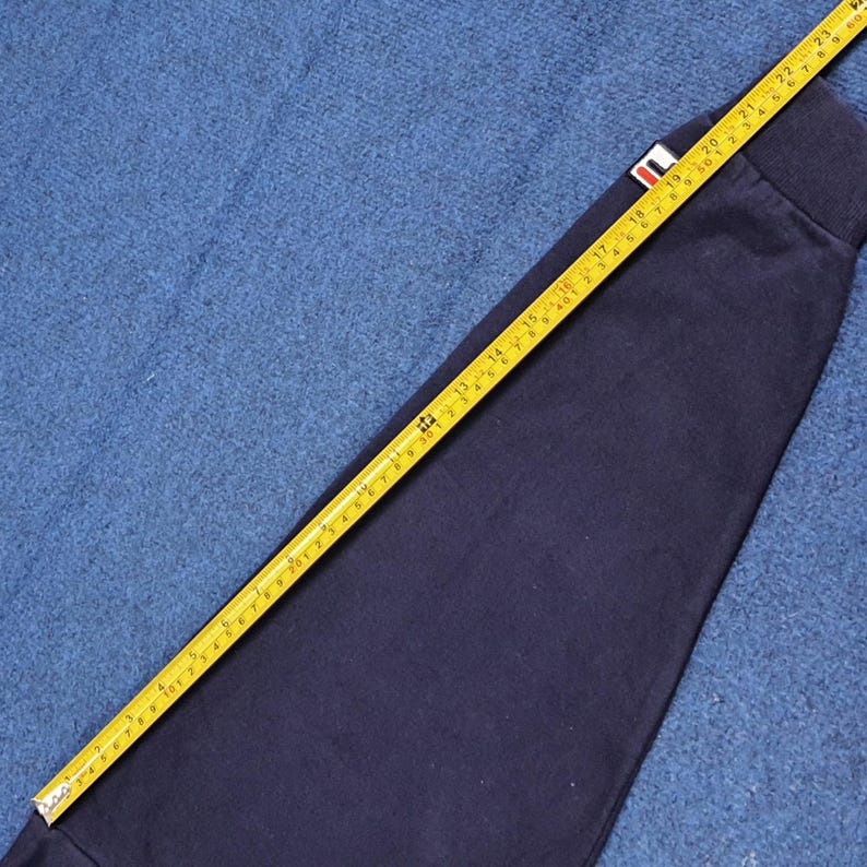 Puede incluir: Manga de sudadera azul marino con un peque&ntilde;o logotipo rojo, blanco y azul. Una cinta m&eacute;trica amarilla se coloca sobre la manga, mostrando medidas en cent&iacute;metros. La manga est&aacute; sobre una superficie azul texturizada.