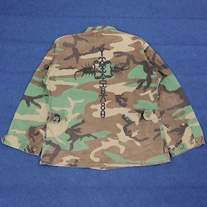 Könnte beinhalten: Tarnmuster-Jacke in Grün-, Braun- und Beigetönen. Auf der Rückseite der Jacke befindet sich ein schwarzes Grafikdesign mit Zahlen und Symbolen. Sie hat lange Ärmel, eine Knopfleiste und zwei Vordertaschen.