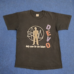 Op de afbeelding: Zwart T-shirt met een zilveren afbeelding van een persoon die in een cirkel staat met een ontwerp van een atoom en een driehoek. De tekst "DEVO" staat in paarse, rode en blauwe letters. De tekst "duty now for the future" staat onder de afbeelding.