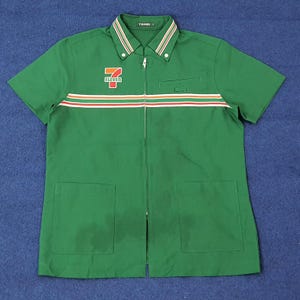 Puede incluir: Camisa de trabajo verde de manga corta con cremallera completa y dos bolsillos delanteros. La camisa presenta una franja roja, blanca y marrón en el pecho y el logotipo de "7-Eleven" en el pecho izquierdo.