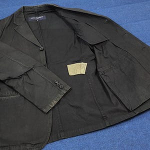 May include: A black blazer with a classic design. The blazer features a notched lapel, three buttons on the sleeve cuffs, and a single-button closure. The interior reveals a pocket and a tag. The blazer is made of a durable fabric.