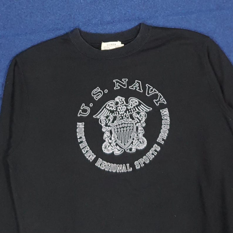 K&ouml;nnte beinhalten: Ein schwarzes Langarmshirt mit einem wei&szlig;en Aufdruck des Emblems des U.S. Navy Northern Regional Sports Program. Das Emblem enth&auml;lt Text und ein Wappen-Design. Das Shirt hat einen Rundhalsausschnitt.