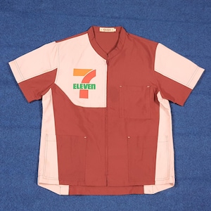 Puede incluir: Una camisa de uniforme de manga corta con un diseño de bloques de color. La camisa presenta una combinación de colores blanco y granate, con el logotipo "7-Eleven" en verde, naranja y rojo en la parte delantera. Tiene dos bolsillos delanteros y cuello mandarín.