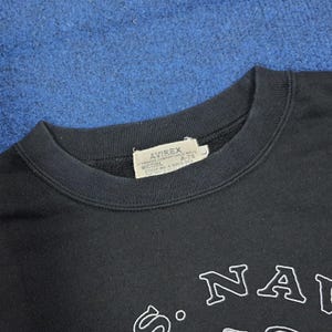 K&ouml;nnte beinhalten: Schwarzes Sweatshirt mit Rundhalsausschnitt und wei&szlig;em Schriftzug &bdquo;NA&ldquo;. Das Sweatshirt hat ein Etikett mit der Aufschrift &bdquo;AVIREX&ldquo;. Das Kleidungsst&uuml;ck besteht aus einem weichen Material.