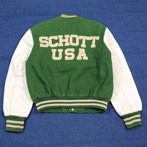 May include: Green and white varsity jacket with the text "SCHOTT USA" on the back. The jacket features green wool fabric on the body and white leather sleeves. Ribbed cuffs and waistband with beige stripes complete the design.
