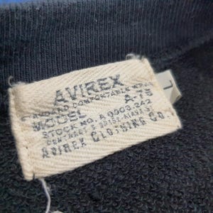 K&ouml;nnte beinhalten: Nahaufnahme eines gewebten Etiketts von einem AVIREX-Kleidungsst&uuml;ck. Das rechteckige Etikett zeigt den Markennamen in fetter Schrift. Der Text enth&auml;lt "STANDARD COMFORTABLE WEAR", "MODEL A-75" sowie Lager- und Vertragsnummern. Der Hintergrund ist ein dunkelblauer Stoff.