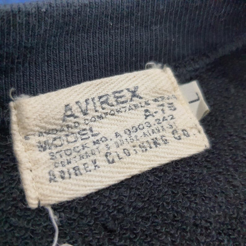 K&ouml;nnte beinhalten: Nahaufnahme eines gewebten Etiketts von einem AVIREX-Kleidungsst&uuml;ck. Das rechteckige Etikett zeigt den Markennamen in fetter Schrift. Der Text enth&auml;lt "STANDARD COMFORTABLE WEAR", "MODEL A-75" sowie Lager- und Vertragsnummern. Der Hintergrund ist ein dunkelblauer Stoff.