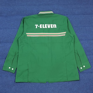 Puede incluir: Camisa de trabajo verde de manga larga con el logo "7-ELEVEN" en blanco en la espalda. La camisa tiene un cuello a rayas verdes y blancas y una franja horizontal en rojo, blanco y naranja. Los puños tienen dos botones blancos cada uno.