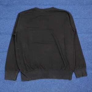 K&ouml;nnte beinhalten: Ein schwarzes Sweatshirt mit Rundhalsausschnitt und langen &Auml;rmeln. Das Sweatshirt hat einen gerippten Saum und B&uuml;ndchen. Der Stoff scheint ein weiches, bequemes Material zu sein. Das Kleidungsst&uuml;ck wird auf einer blauen Oberfl&auml;che pr&auml;sentiert.