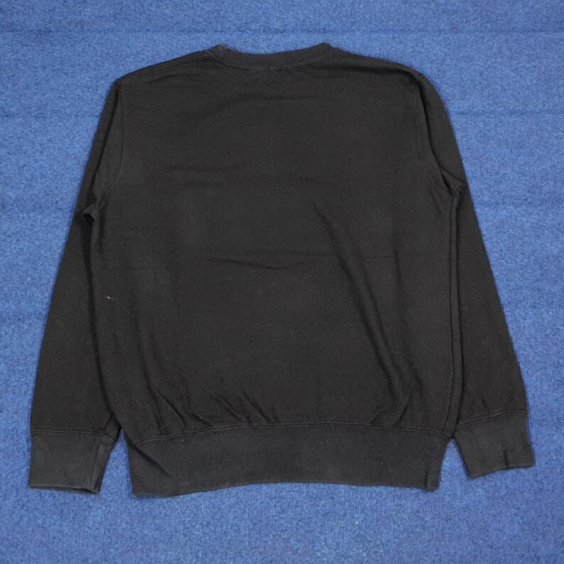 K&ouml;nnte beinhalten: Ein schwarzes Sweatshirt mit Rundhalsausschnitt und langen &Auml;rmeln. Das Sweatshirt hat einen gerippten Saum und B&uuml;ndchen. Der Stoff scheint ein weiches, bequemes Material zu sein. Das Kleidungsst&uuml;ck wird auf einer blauen Oberfl&auml;che pr&auml;sentiert.