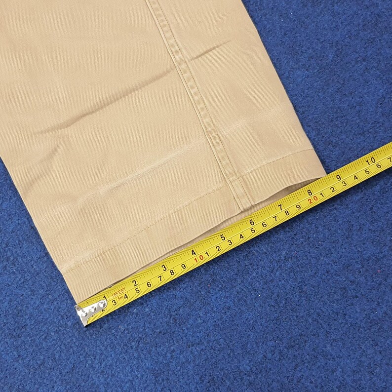 May include: Tan-colored fabric with a seam detail is measured with a yellow tape measure. The tape measure shows a measurement of approximately 3 inches. The fabric appears to be part of a pair of pants.