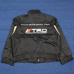 May include: Black Toyota Motor Sports TRD jacket with white and beige detailing. The back of the jacket displays the TRD logo and the text "TOYOTA RACING DEVELOPMENT". The jacket has a stand-up collar and a button closure at the waist.