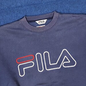 Puede incluir: Sudadera azul marino con el nombre de la marca "FILA" en letras blancas grandes con contorno rojo. La sudadera tiene cuello redondo y una peque&ntilde;a etiqueta de la marca en el cuello.