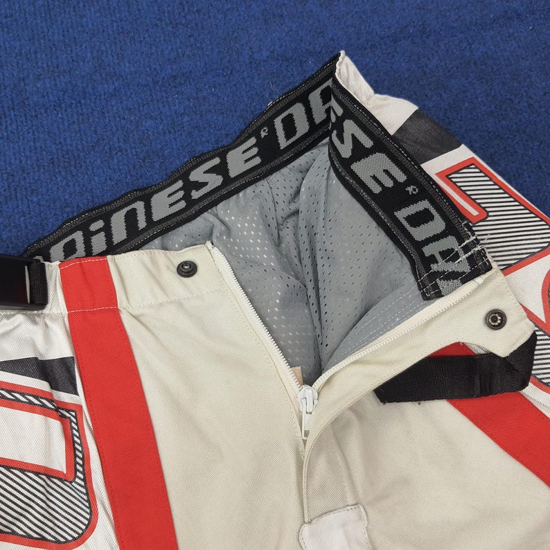 Puede incluir: Primer plano de pantalones de moto en crema, rojo y negro. Los pantalones tienen cremallera, corchetes y forro de malla. La cintura lleva el nombre de la marca en letras negras.