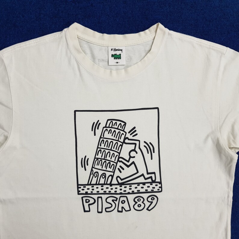 Puede incluir: Camiseta blanca con un gr&aacute;fico en blanco y negro de una persona empujando la Torre Inclinada de Pisa. El gr&aacute;fico est&aacute; en un marco cuadrado con el texto "PISA 89" debajo.