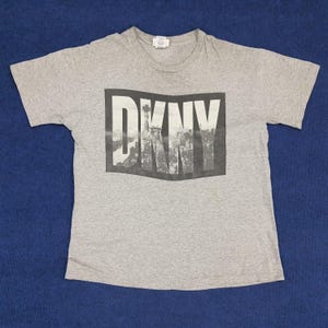 May include: Grey short-sleeved t-shirt with a black and white graphic print of the word "DKNY" with a cityscape in the background.