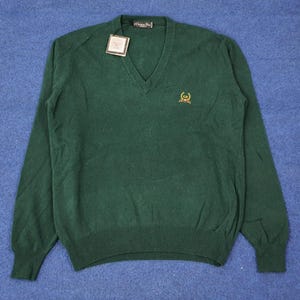 Può includere: Un maglione verde scuro con scollo a V e maniche lunghe. Il maglione ha orlo e polsini a coste e un piccolo logo ricamato sul petto. Un'etichetta è attaccata vicino allo scollo. Il maglione è in materiale morbido.