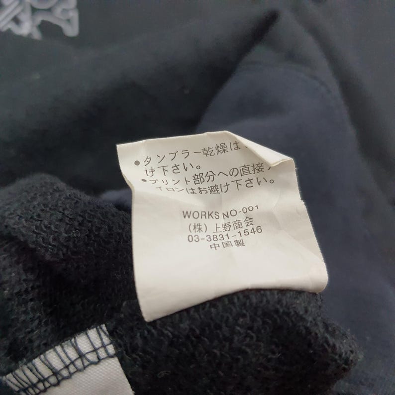 K&ouml;nnte beinhalten: Nahaufnahme eines dunkelmarineblauen Kleidungsst&uuml;cks mit einem wei&szlig;en Etikett. Das Etikett enth&auml;lt japanischen Text und den Text "WORKS NO-001". Der Stoff des Kleidungsst&uuml;cks ist im Vordergrund sichtbar.