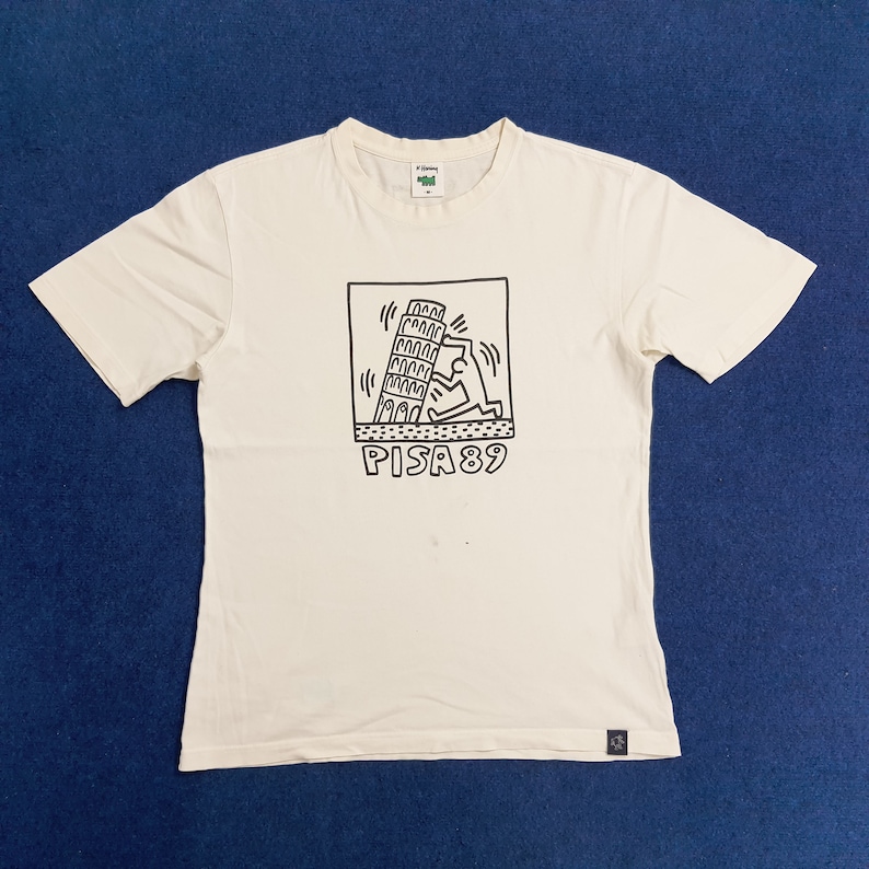 Puede incluir: Una camiseta blanca con un gr&aacute;fico en blanco y negro de una persona empujando la Torre Inclinada de Pisa. El gr&aacute;fico est&aacute; rodeado de un cuadrado negro con el texto "PISA 89" debajo.