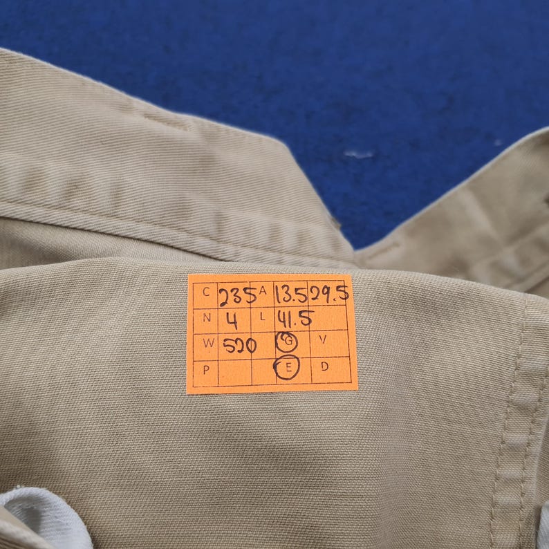 May include: Close-up of a tan-colored garment with a collar and buttoned placket. An orange tag with black text is attached, displaying numbers and letters. The fabric appears to be a sturdy, woven material.