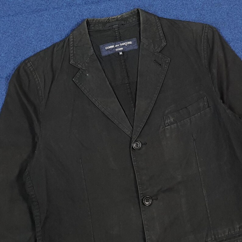 May include: A black blazer with a notched lapel, two-button closure, and a single chest pocket. The blazer has a tailored fit and a label that reads "COMME des GAR&Ccedil;ONS HOMME".
