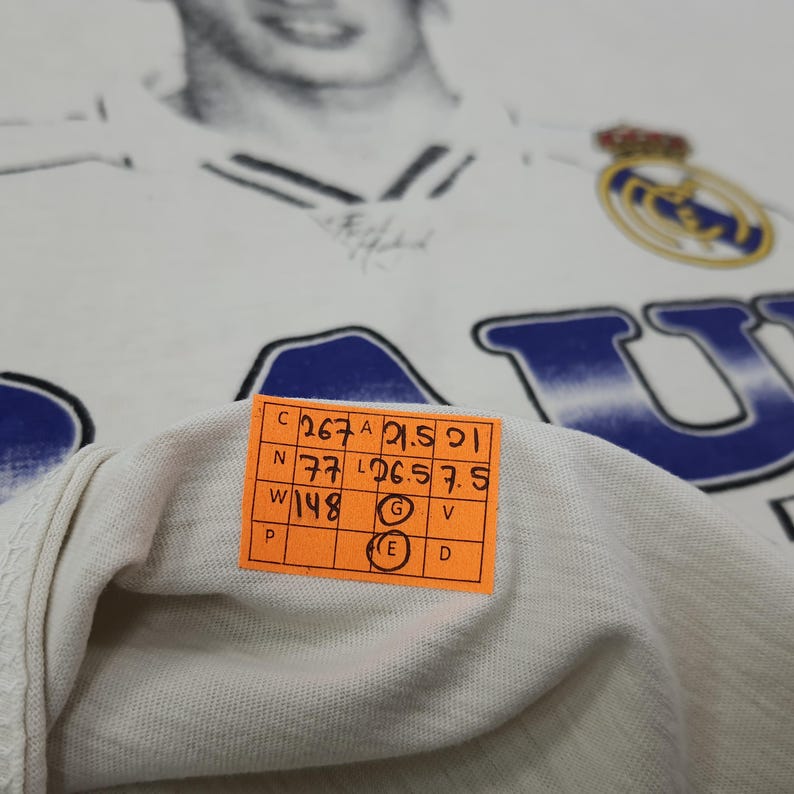 May include: White t-shirt featuring a faded image of a soccer player and the Real Madrid logo. The shirt has blue lettering and a small orange tag with measurements. The shirt is made of a soft, likely cotton material.