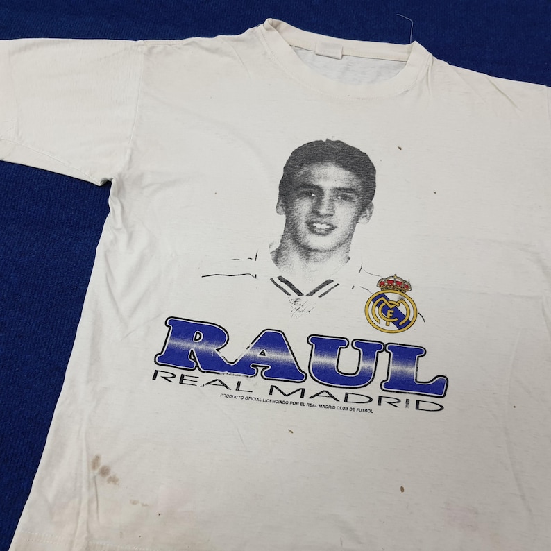 May include: White t-shirt featuring a black and white portrait of a young man, with the name "RAUL" in blue block letters. The Real Madrid logo is also visible. The shirt is slightly wrinkled and has some stains.