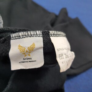 K&ouml;nnte beinhalten: Nahaufnahme eines schwarzen Kleidungsst&uuml;cketiketts mit dem Avirex-Logo, einem goldenen Emblem aus Fl&uuml;geln und einem Stern. Auf dem Etikett steht "Avirex AUTHENTIC PRODUCT". Ein zweites Etikett zeigt Pflegehinweise und japanischen Text.