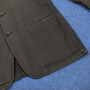 May include: Close-up of a dark gray blazer with two buttons and a pocket. The blazer is made of a textured fabric and has a classic design. The buttons are black and round.