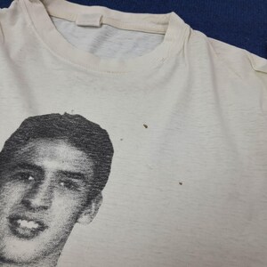 May include: Off-white t-shirt featuring a black and white portrait of a person. The shirt has a crew neck and appears to be vintage. The image is a close-up of the shirt's front, showing the graphic and fabric texture.