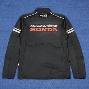 May include: Black zip-up jacket with white and gray accents. The jacket features the text "MUGEN 無限 HONDA" in red and white. The jacket has a small logo on the upper back and white stripes on the sleeves.