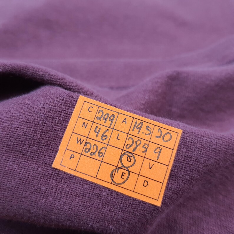 May include: Close-up of a textured, deep purple fabric. An orange tag with black handwritten numbers and letters is attached to the fabric. The tag has the letters C, N, W, A, L, P, G, E, V, and D.
