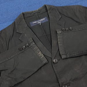 May include: A black blazer with a notched lapel, three-button cuffs, and a two-button closure. The blazer has a label that reads "Comme des Garcons Homme". The jacket is made of a durable fabric.