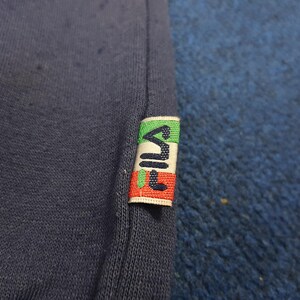 Puede incluir: Primer plano de una tela azul marino con una etiqueta de la marca Fila. La etiqueta presenta el nombre de la marca en blanco, con rayas verdes, rojas y blancas. La tela tiene un aspecto ligeramente texturizado.