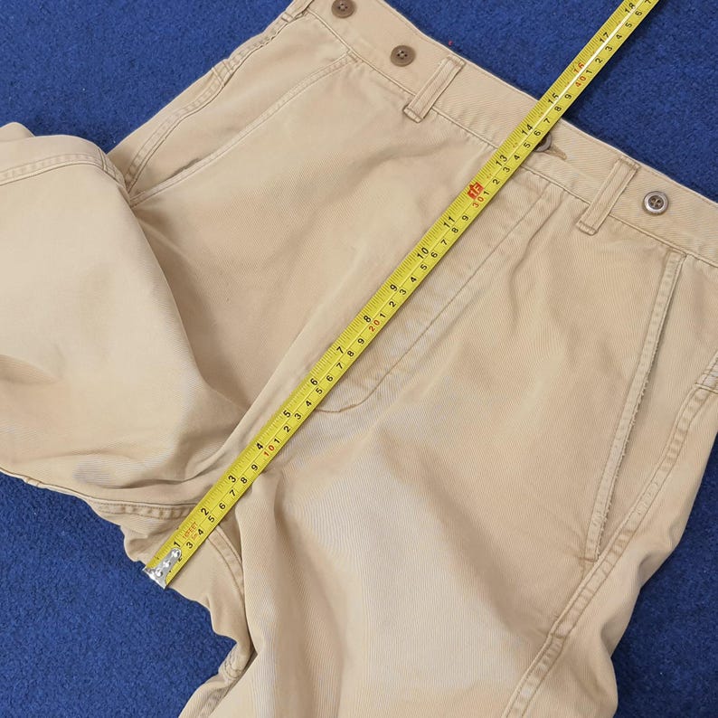 May include: Khaki-colored pants with a straight leg design. The pants have a button and zipper closure, belt loops, and pockets. A yellow measuring tape is laid across the pants, showing measurements in inches. The pants are folded on a blue surface.