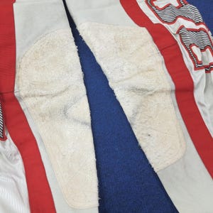 Puede incluir: Primer plano de un pantal&oacute;n de moto gris claro con rayas rojas y parches blancos. El pantal&oacute;n tiene rodilleras blancas y un dise&ntilde;o blanco y rojo en el lateral.