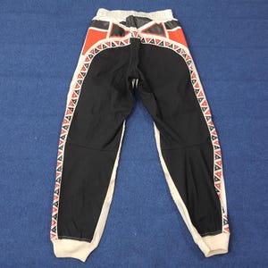 Puede incluir: Pantalones deportivos negros con detalles geom&eacute;tricos blancos y rojos. Los pantalones tienen una franja blanca en los laterales y una cintura blanca. La parte trasera de los pantalones tiene un dise&ntilde;o rojo y negro.